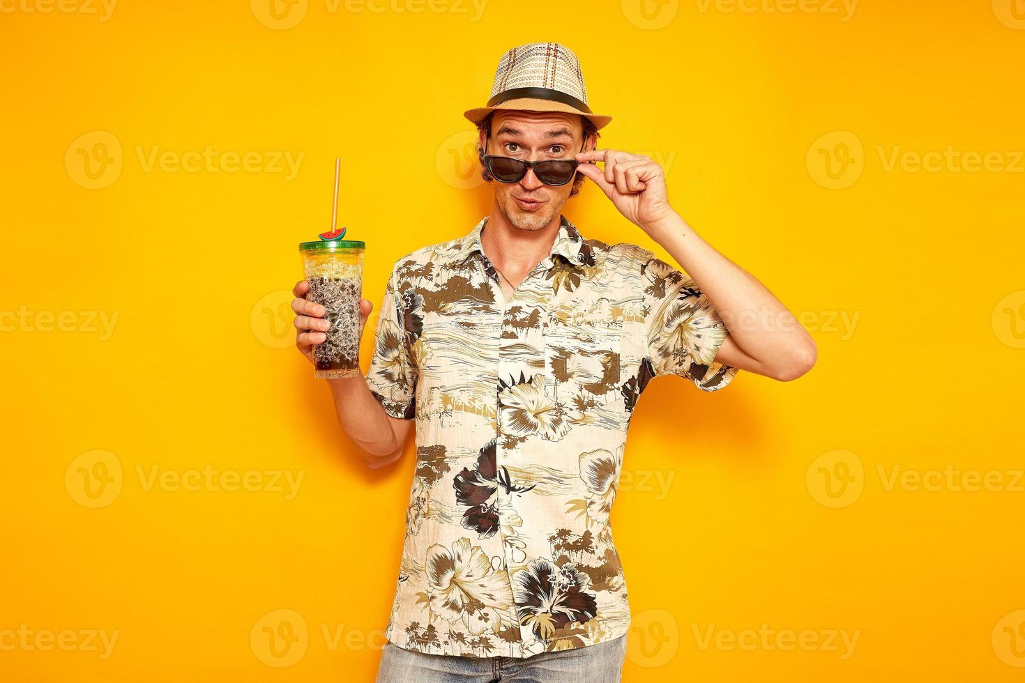 pleasantly surprised face, male tourist traveler in hat, Hawaiian shirt on vacation with drink in cocktail glass lowered his sunglasses with his hand. isolated on yellow background with space for text photo