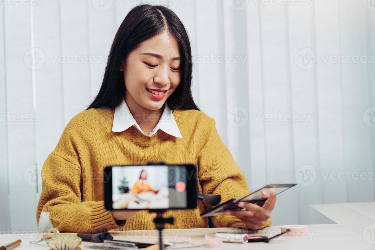 Beautiful asian woman blogger using camera phone recording vlog video live streaming and showing how to make up cosmetics at home. photo