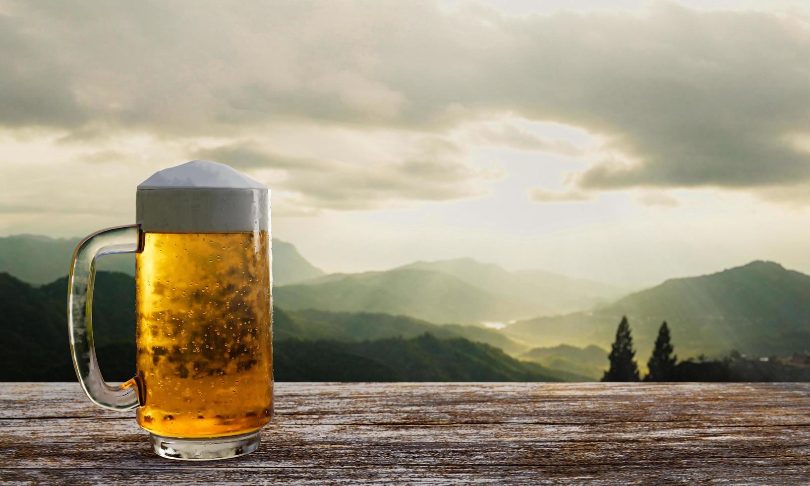 cerveza de barril o artesanal en vidrio transparente con espuma de cerveza en la parte superior puesta en un juego de mesas y sillas de madera. asientos de salón en la terraza de madera con fondo de montaña con luz de dios o luz solar.representación 3d foto