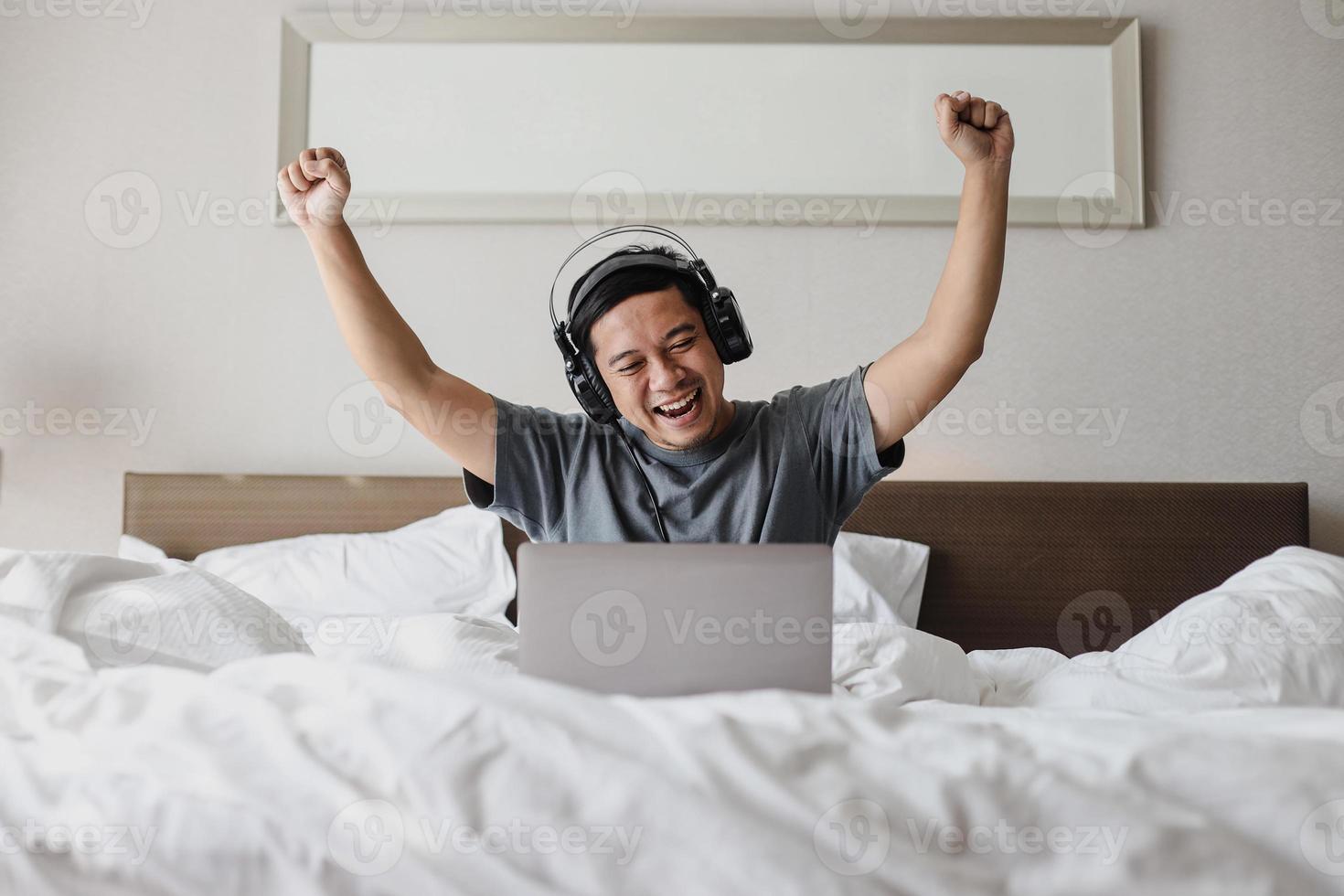 hombre asiático feliz en auriculares mirando a la computadora portátil con emocionado expresando gesto ganador en la cama foto