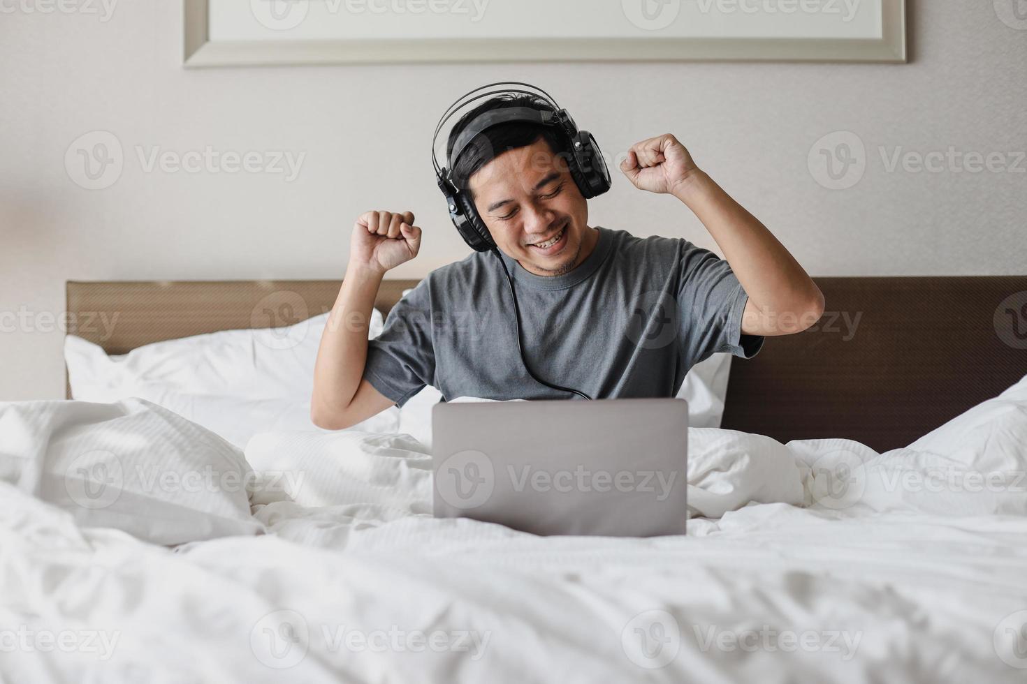 Smiling Asian Man listening music and dancing with headphones and laptop on the bed photo