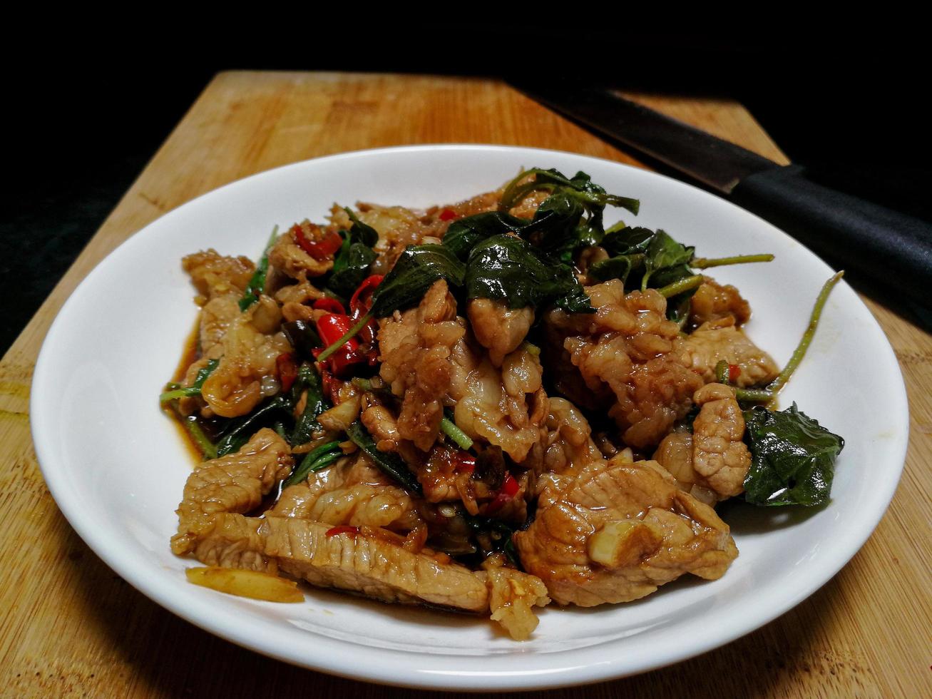 Stir fried beef with holy basil in a white plate, placed on a wooden chopping board and knife. Popular food in Thailand consists of meat, garlic, fresh chilies and basil leaves. photo