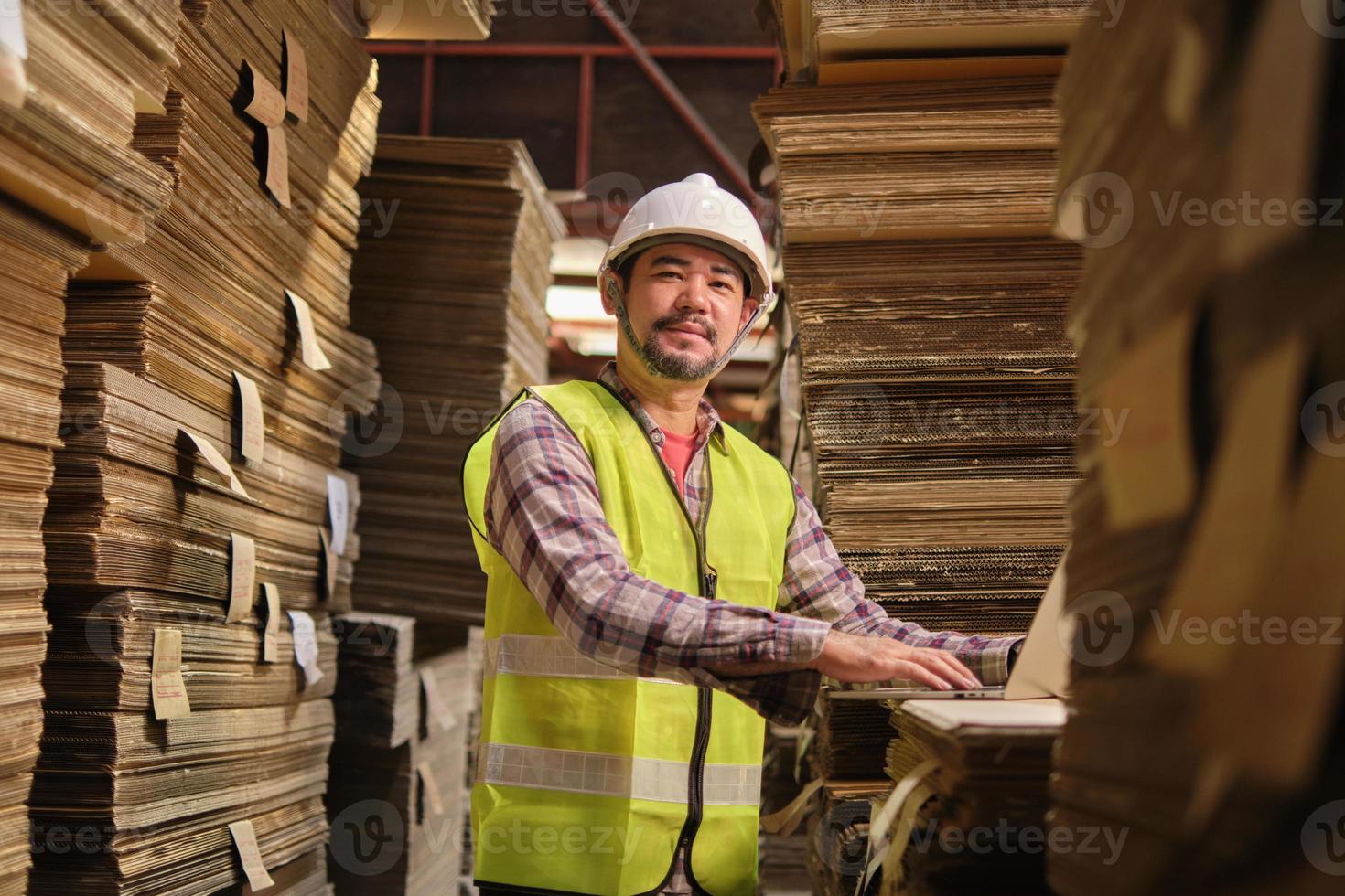 trabajador masculino asiático con uniforme de seguridad y sombrero duro mira la cámara, inspector supervisor con computadora portátil, pedido de existencias en el almacén de la fábrica, pilas de fabricación de cartón apilado, industria de producción. foto