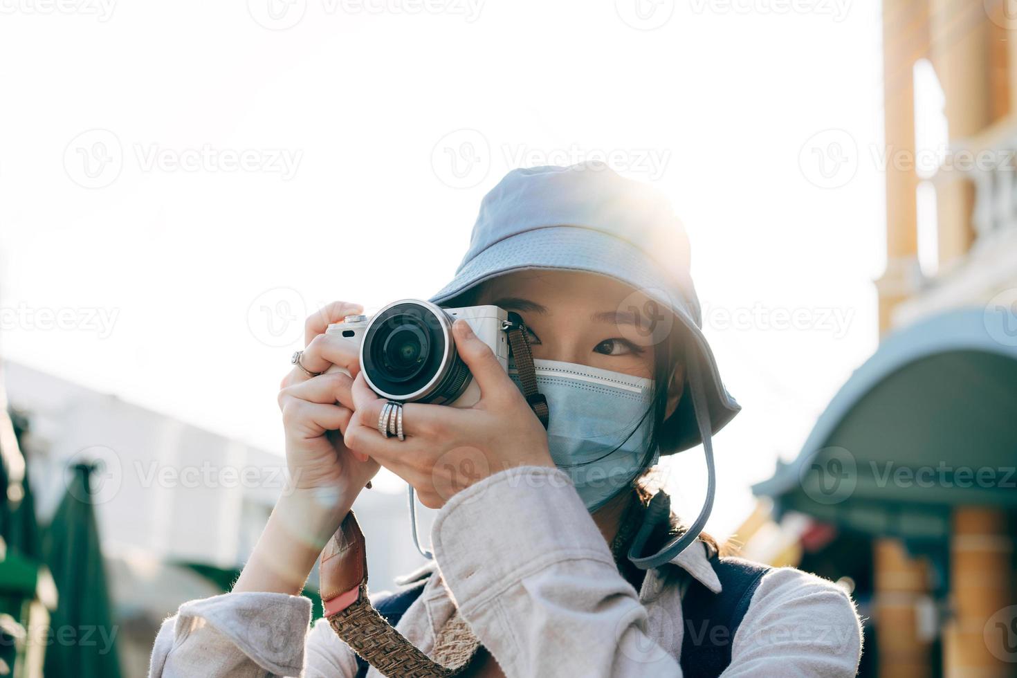 Una joven adulta asiática que viaja con mochila usa mascarilla para tomar una foto con la cámara.