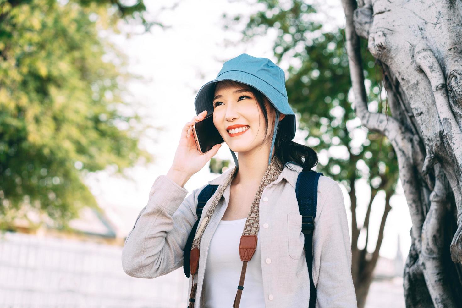 una joven viajera asiática adulta usa un sombrero azul y una mochila usando internet de teléfono móvil para llamar a viajar. foto