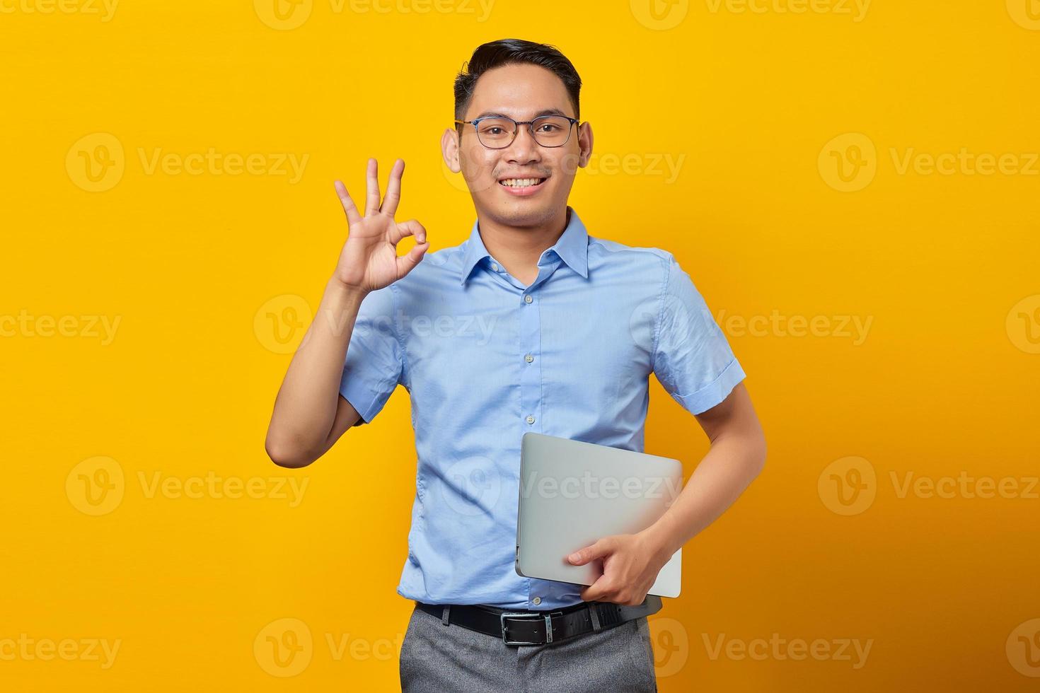 Portrait of smiling handsome Asian man in glasses holding laptop and gesturing okay sign isolated on yellow background. businessman and entrepreneur concept photo