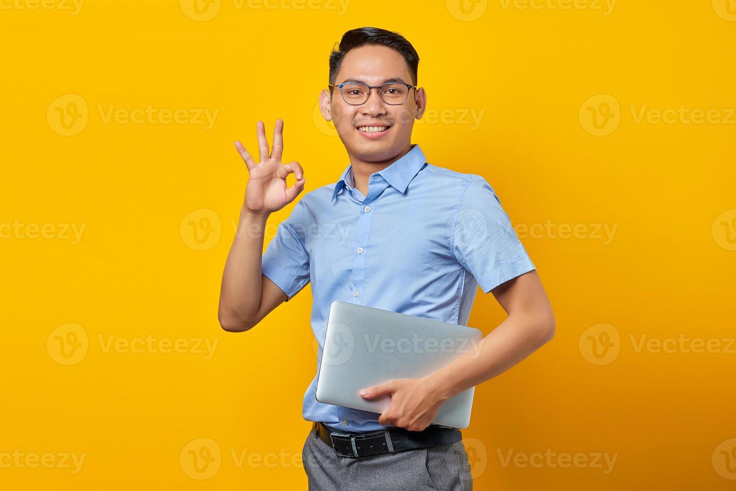 Portrait of smiling handsome Asian man in glasses holding laptop and gesturing okay sign isolated on yellow background. businessman and entrepreneur concept photo