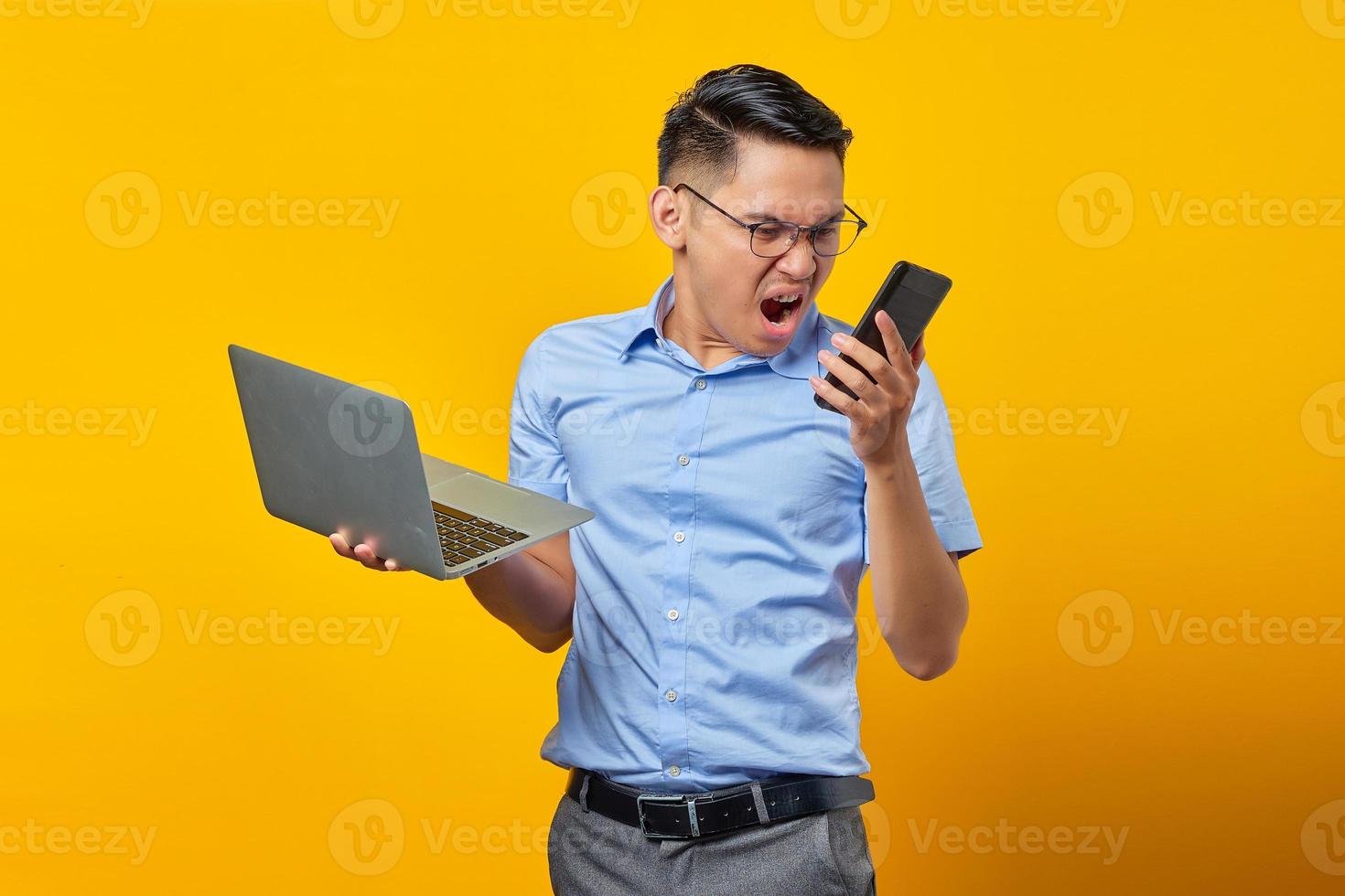 Annoyed young asian man Asian in glasses holding laptop and using a mobile phone while loudly shouting isolated on yellow background. businessman and entrepreneur concept photo