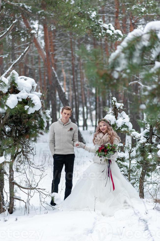 Cheerful bride and groom in beige knitted pullovers are walking in snowy forest. Winter wedding photo