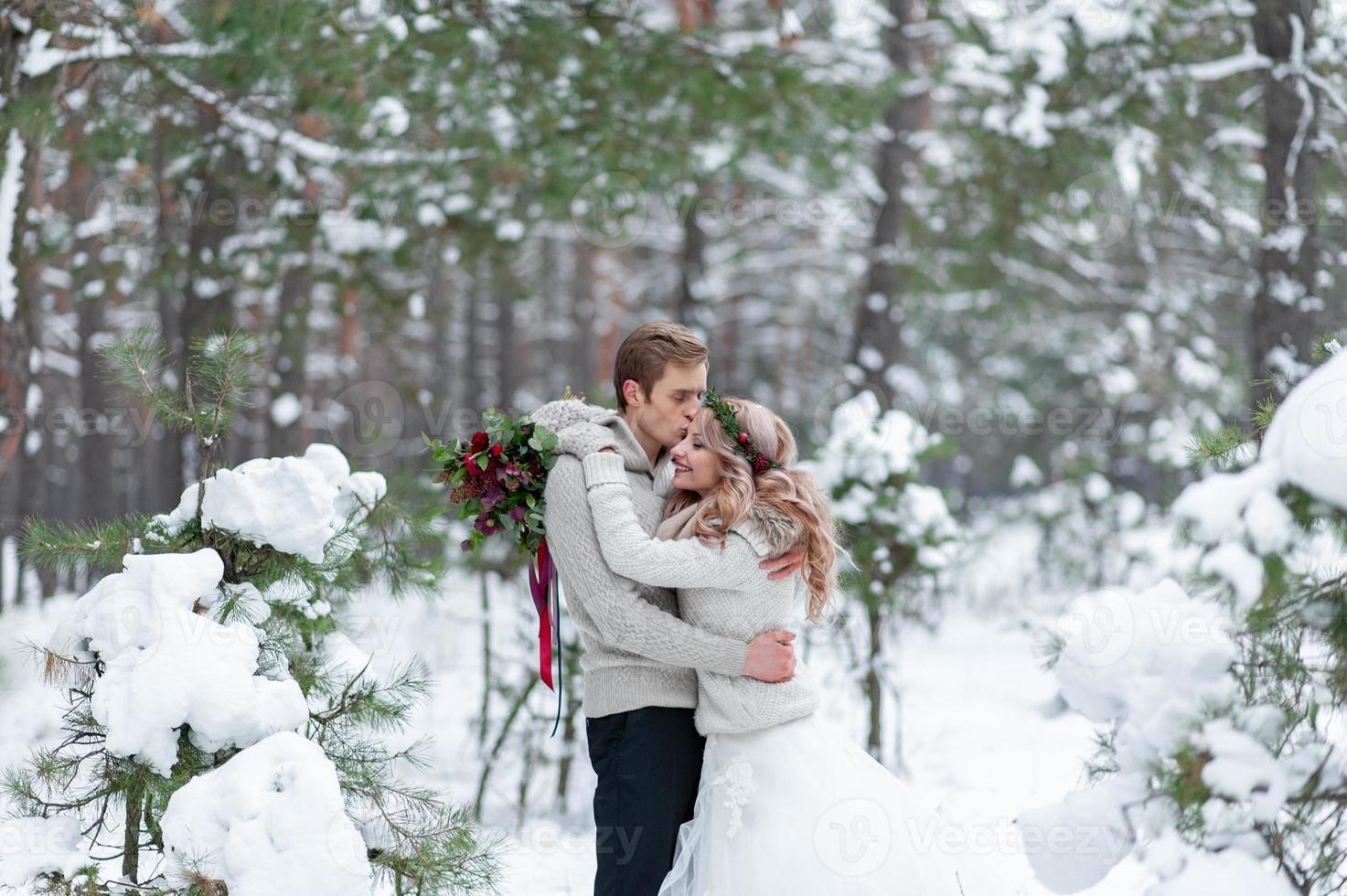 Bride and groom in beige knitted pullovers in snowy forest. Newlyweds is touching foreheads. Winter wedding. Copy space photo