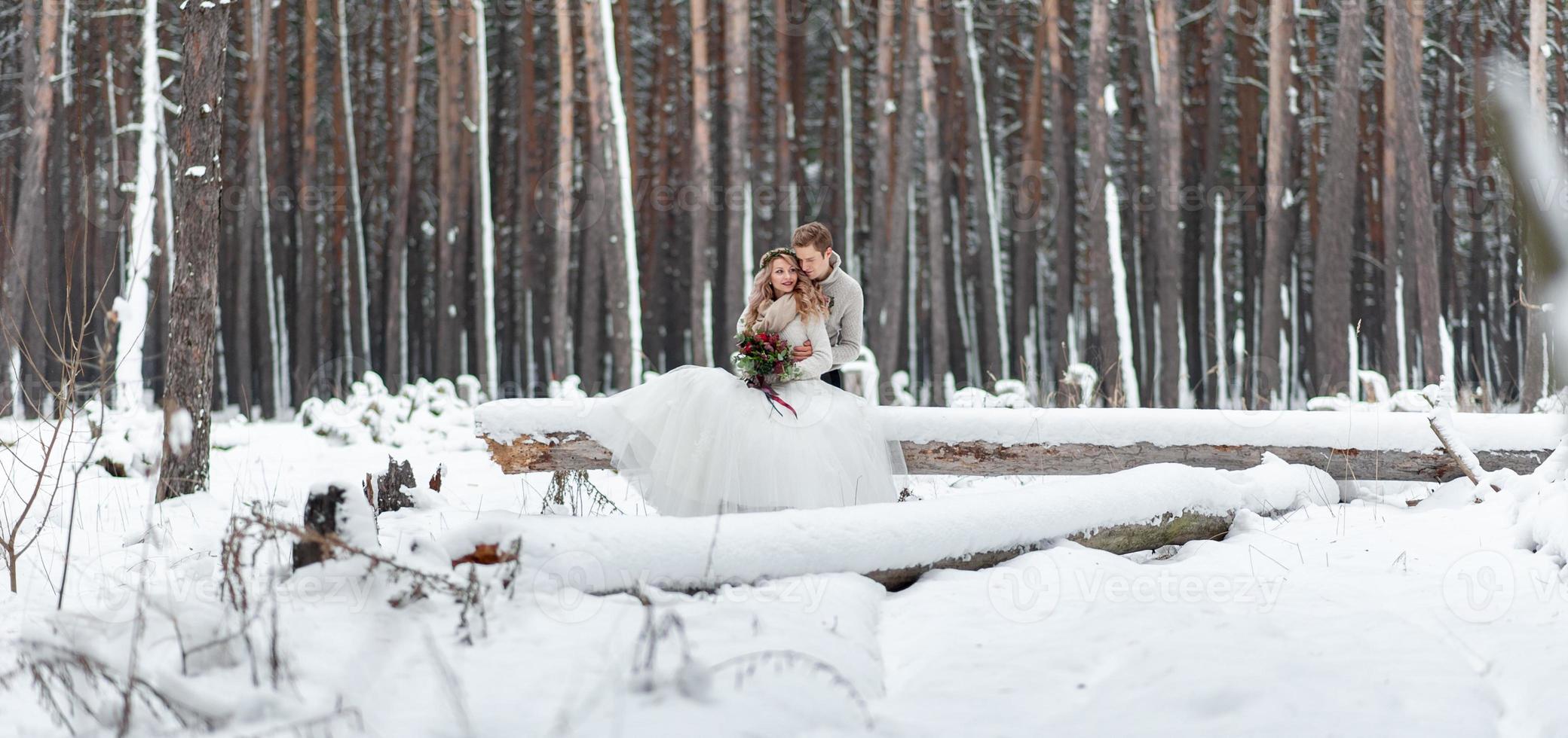 Cute couple in love with a bouquet are sitting on the log on background of the winter forest. Artwork. Winter wedding. Copy space photo