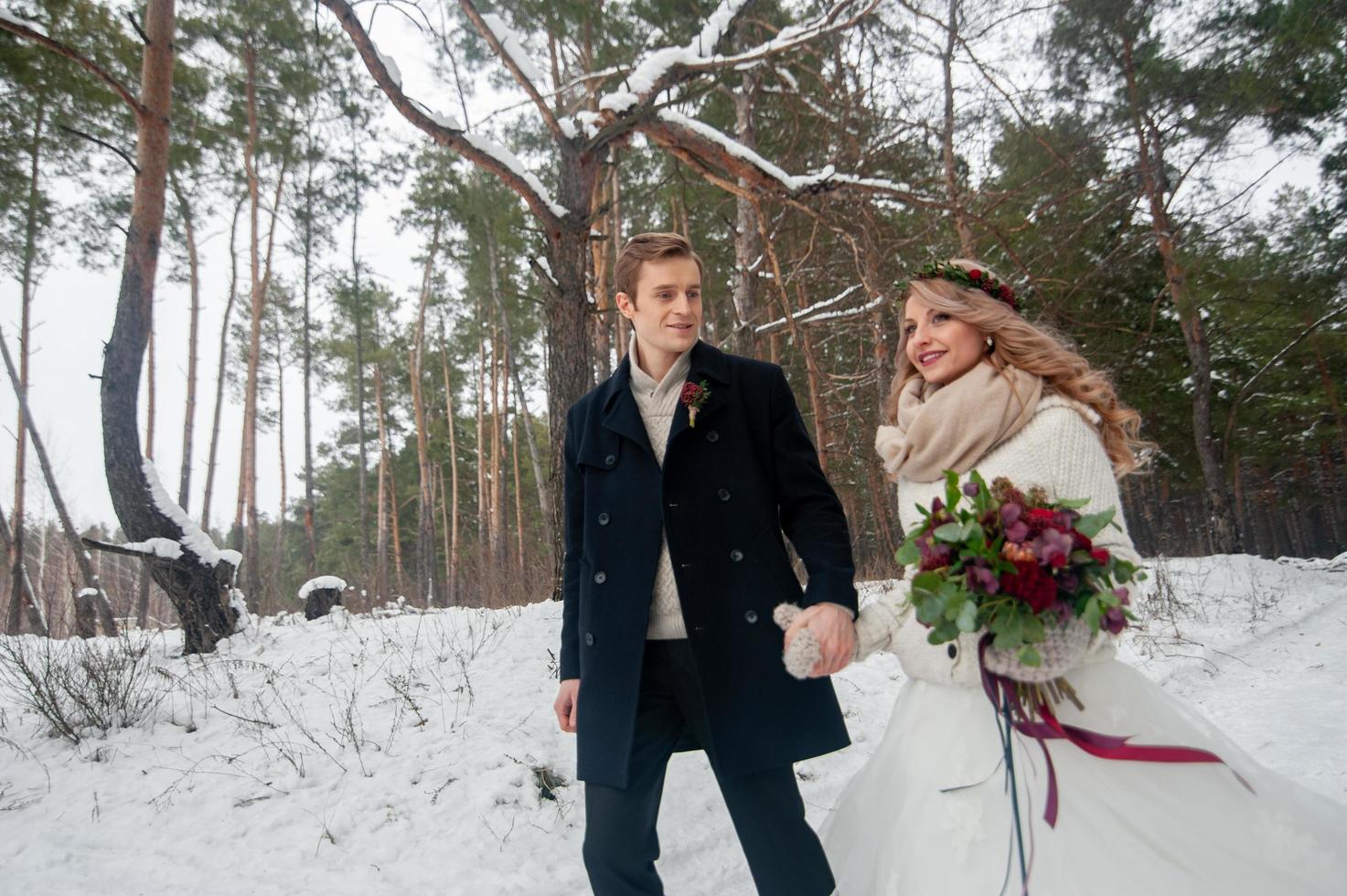 Cheerful bride and groom in beige knitted pullovers are walking in snowy forest. Winter wedding photo