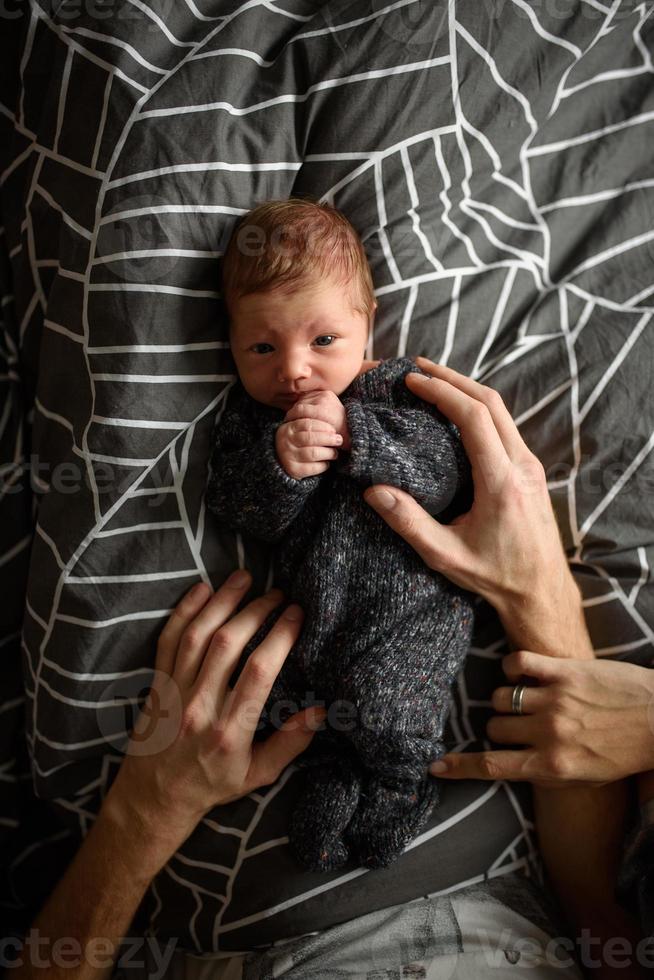 A little newborn boy is looking at the window in his crib. A boy born at 8 months of gestation. There is a knitted suit on the boy. photo