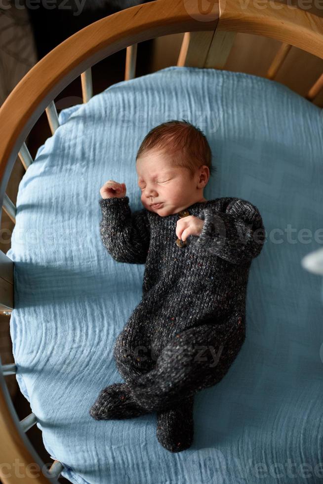 A little newborn boy is looking at the window in his crib. A boy born at 8 months of gestation. There is a knitted suit on the boy. photo