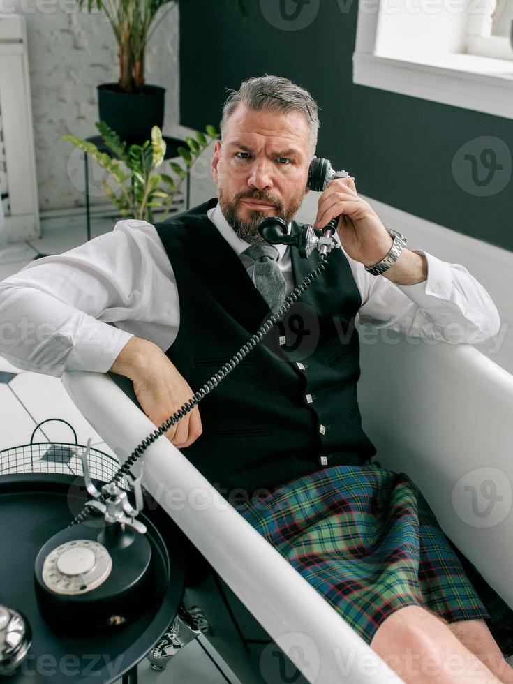 handsome mature courageous stylish man scotsman in kilt and suit talking on the phone from home. Style, fashion, lifestyle, lockdown, culture, ethnic concept. photo