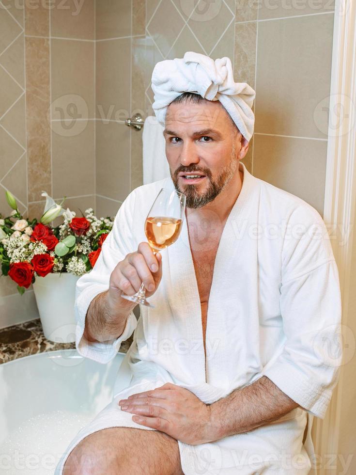 handsome mature man in white bathrobe and towel on his head sitting on bathroom with foam and drinking rose wine. Spa, relax, lifestyle, enjoy life concept. photo