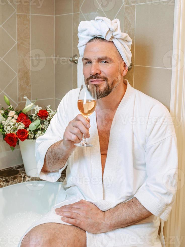 apuesto hombre maduro con albornoz blanco y toalla en la cabeza sentado en el baño con espuma y bebiendo vino rosado. spa, relax, estilo de vida, disfrute del concepto de vida. foto