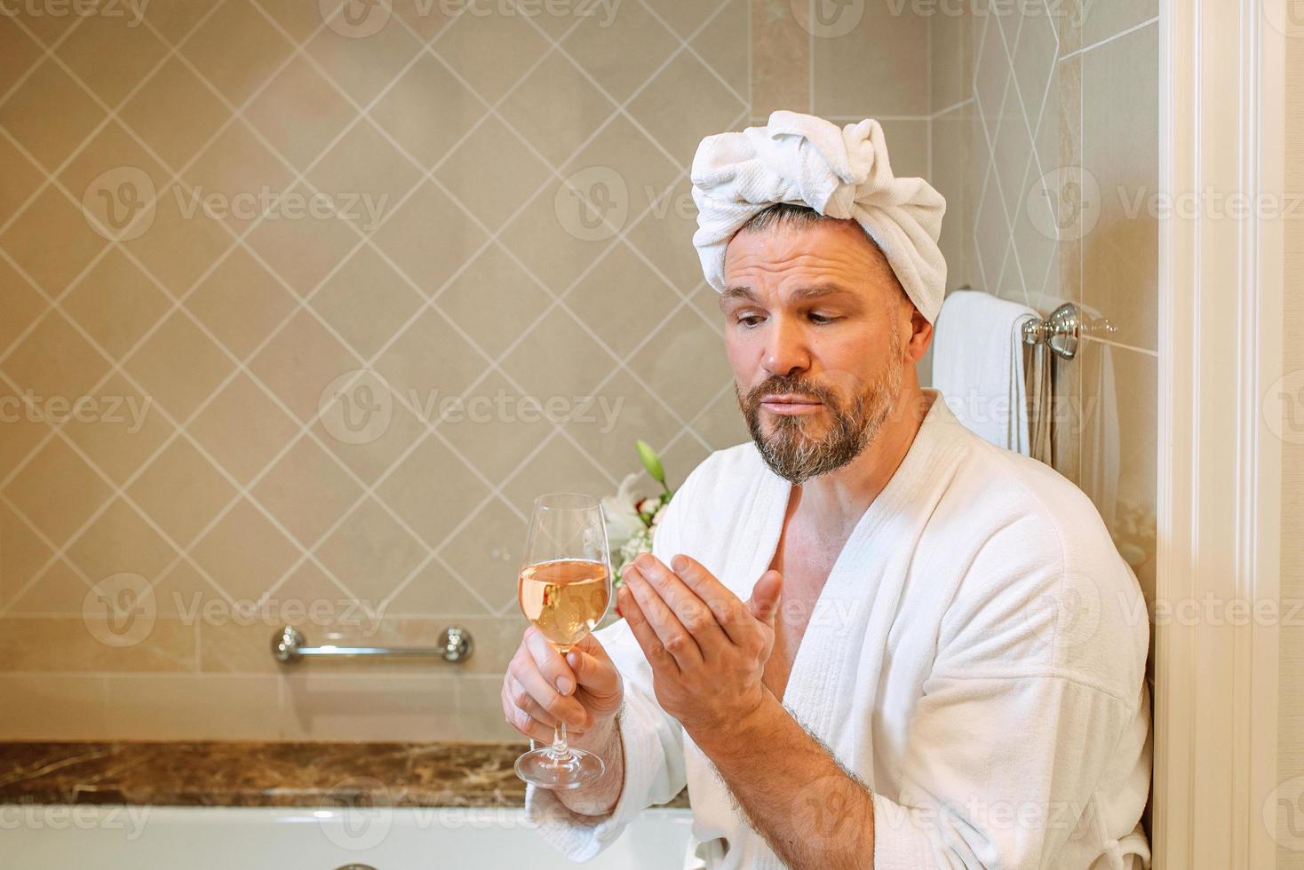 handsome mature man in white bathrobe and towel on his head sitting on bathroom with foam and drinking rose wine. Spa, relax, lifestyle, enjoy life concept. photo