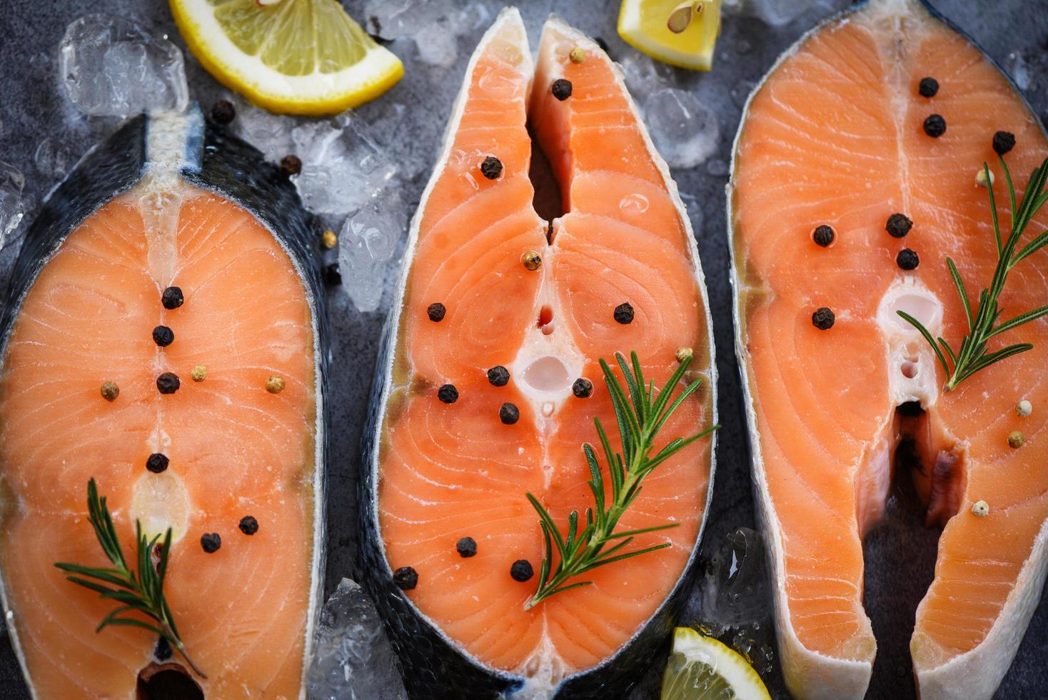 Raw salmon fish fillet and ice for cooked steak seafood - Close up fresh salmon steak with herbs and spices lemon rosemary on black plate background photo