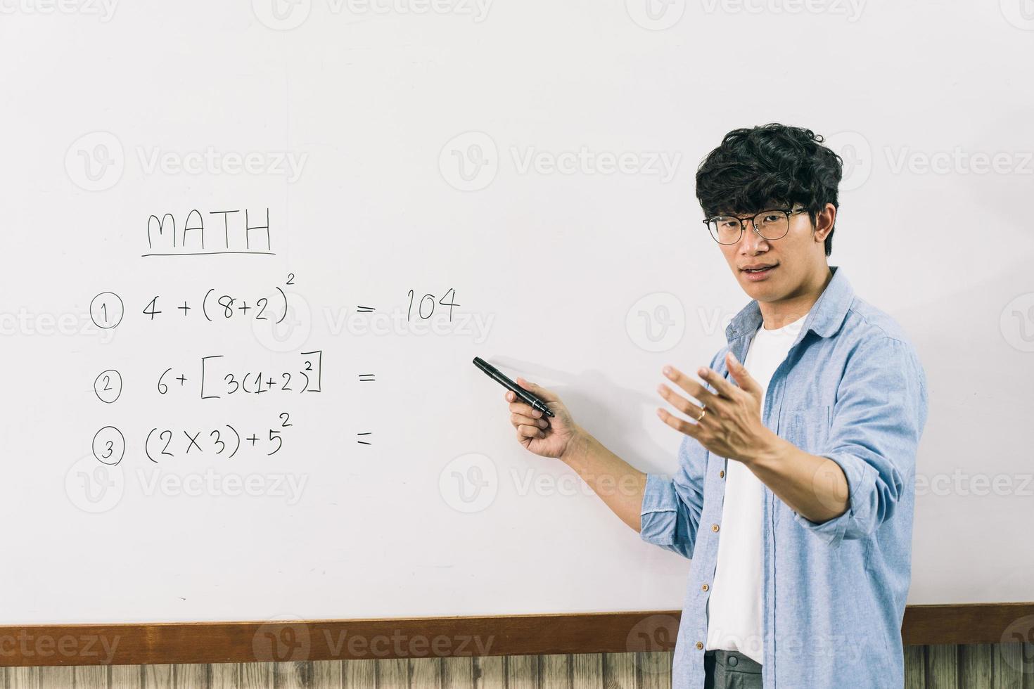 Asian male teacher is teaching students at the classroom while pointing at numbers on the white board. photo