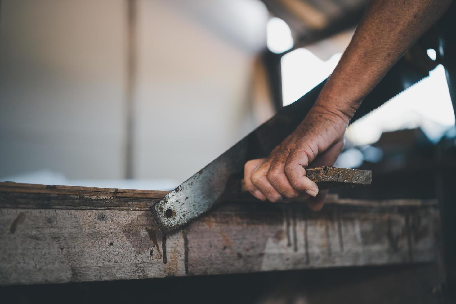 Senior old carpenter using saw working on wood craft at workshop to produce construction material or wooden furniture. photo