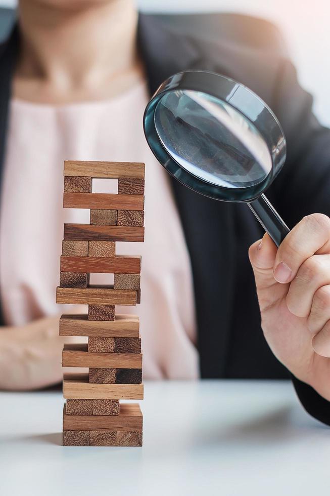 mano de empresaria con lupa de vidrio sobre la torre de bloques de madera. análisis de negocios, gestión de riesgos, estudio, oportunidades y conceptos de estrategia foto