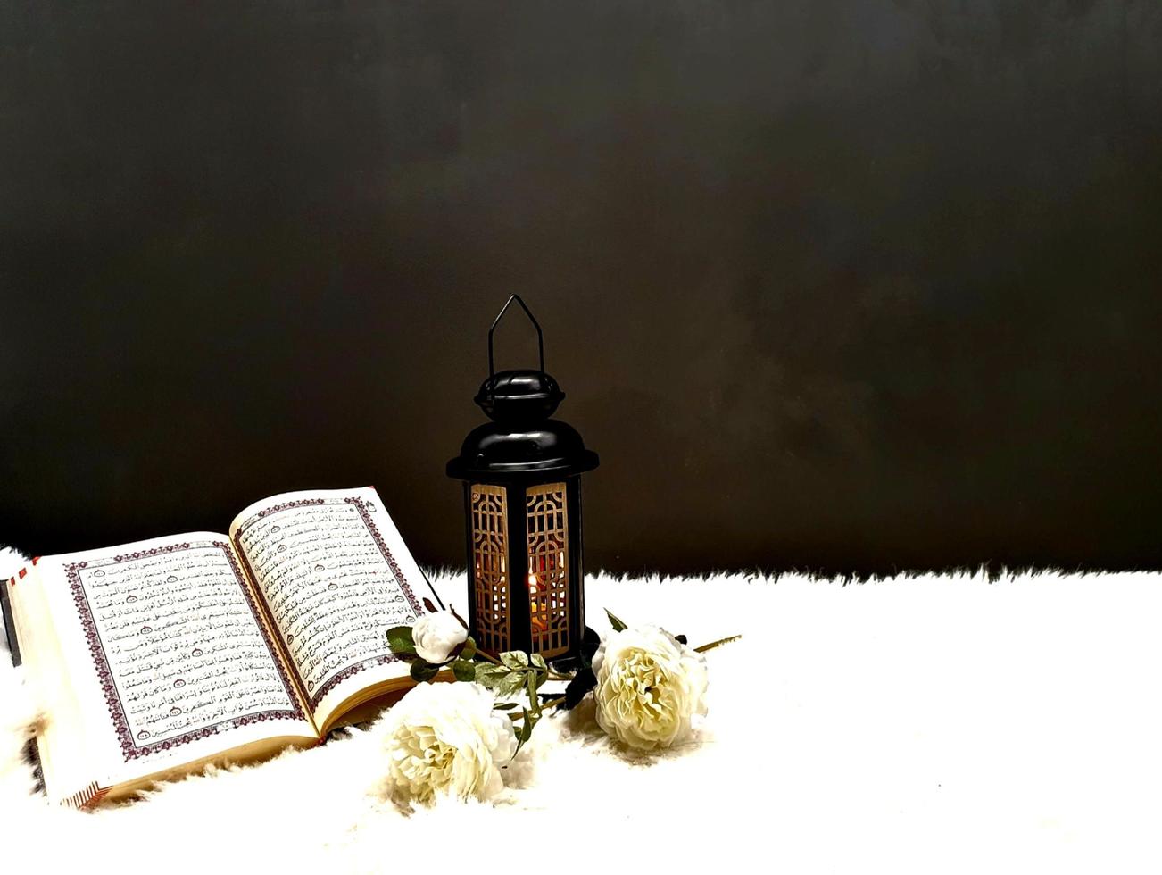 The lantern of Ramadan is black in color, luminous, decorated with wooden motifs, next to the Holy Quran, with a few white roses photo