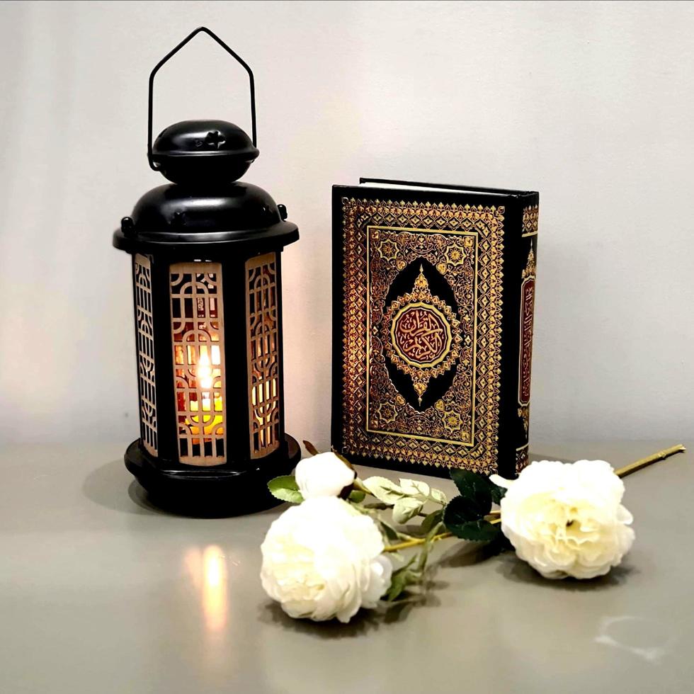 The lantern of Ramadan is black in color, luminous, decorated with wooden motifs, next to the Holy Quran, with a few white roses photo