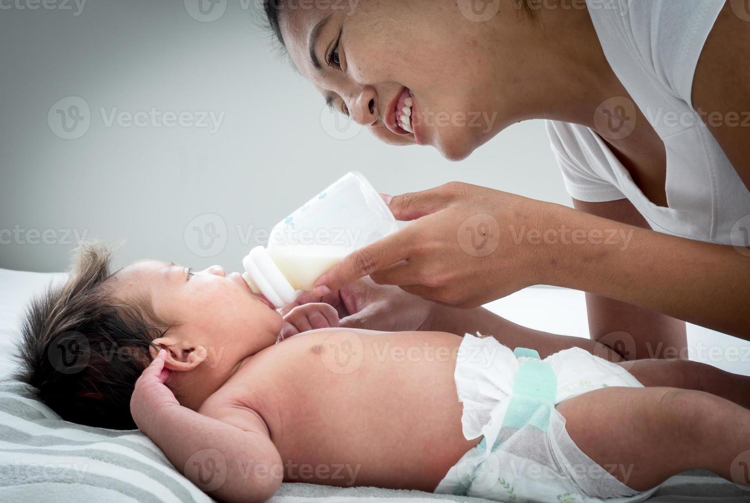 Portrait of Asian mother feeds the Australian Asian three weeks newborn with milk formula from a bottle, concept of motherhood and childhood or infant lying on the white bed. photo