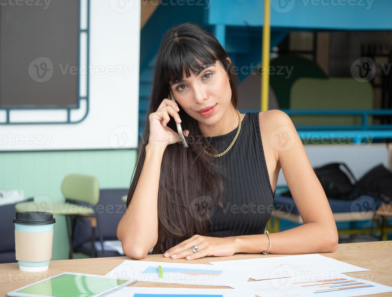 atractiva mujer de negocios caucásica que usa un teléfono móvil y hace una llamada mientras se sienta en la mesa con un documento de información y trabaja en una oficina moderna foto