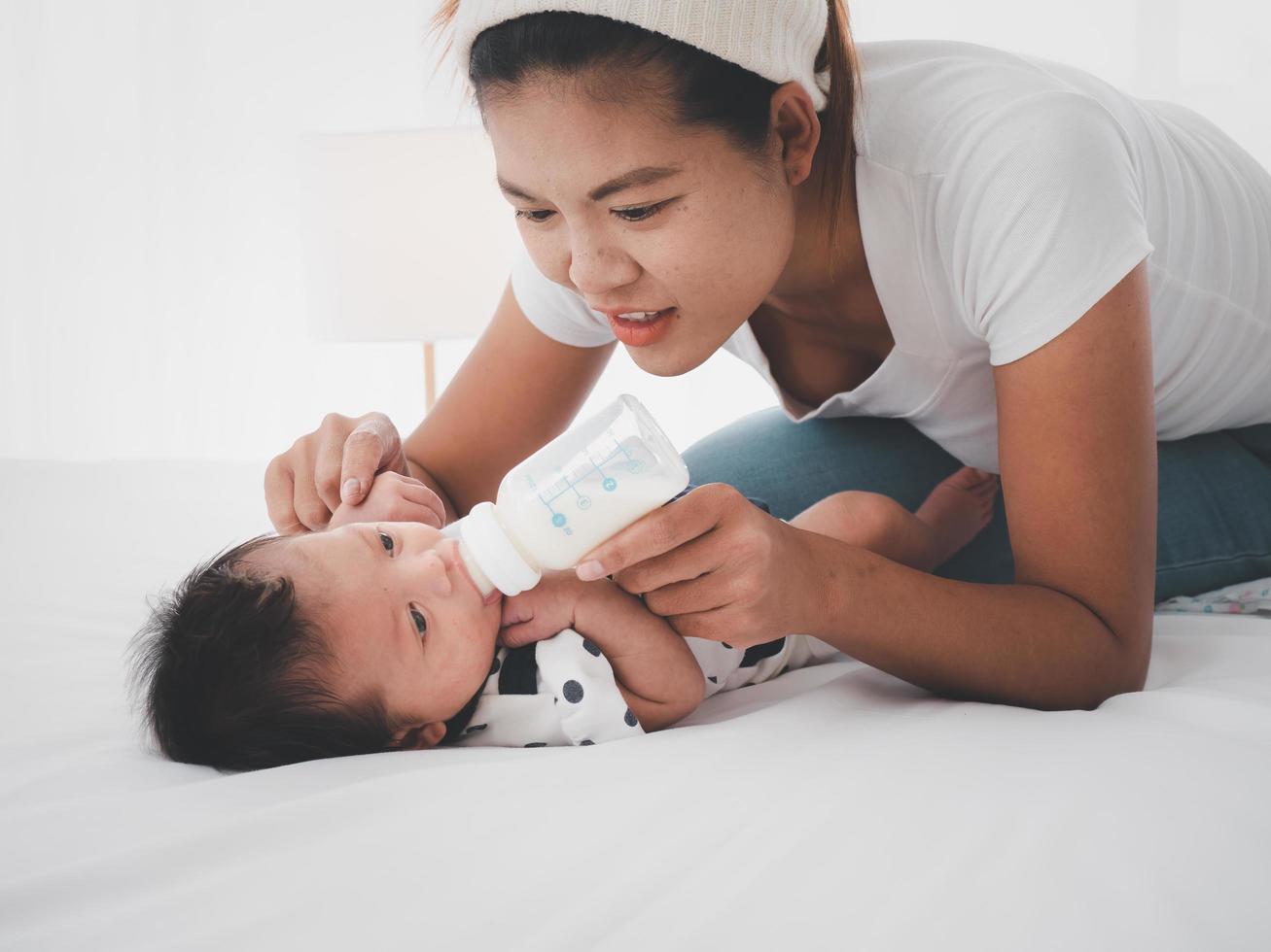 el retrato de una madre asiática alimenta al recién nacido asiático australiano de tres semanas con fórmula láctea de un biberón, concepto de maternidad e infancia o bebé acostado en la cama blanca. foto