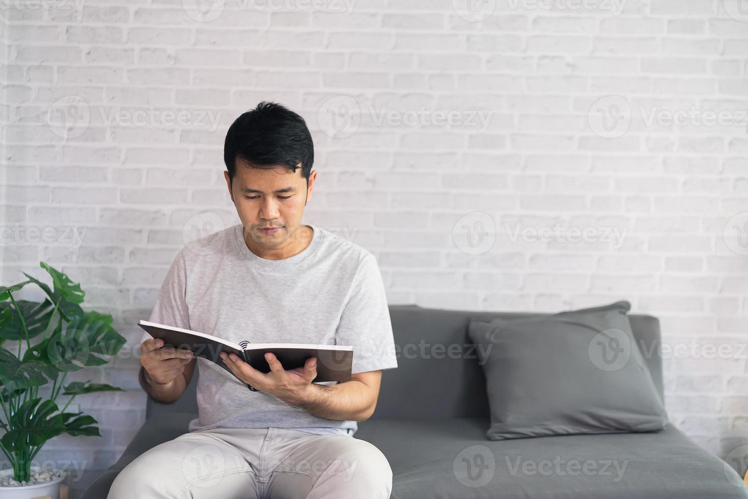 Portrait of joyful asian man reading book while sitting on floor in his living room. Asian man relex reading book smile at sofa In the house. photo