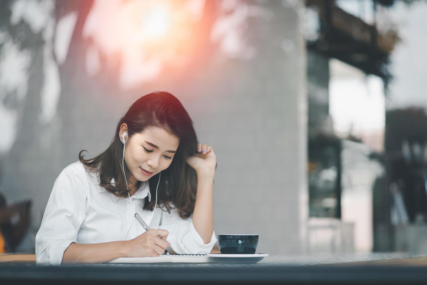 Asian woman sitting and using a pen to write on a paper notebook with a laptop computer and wearing earphones. Work from anywhere concept. Work from home concept. photo
