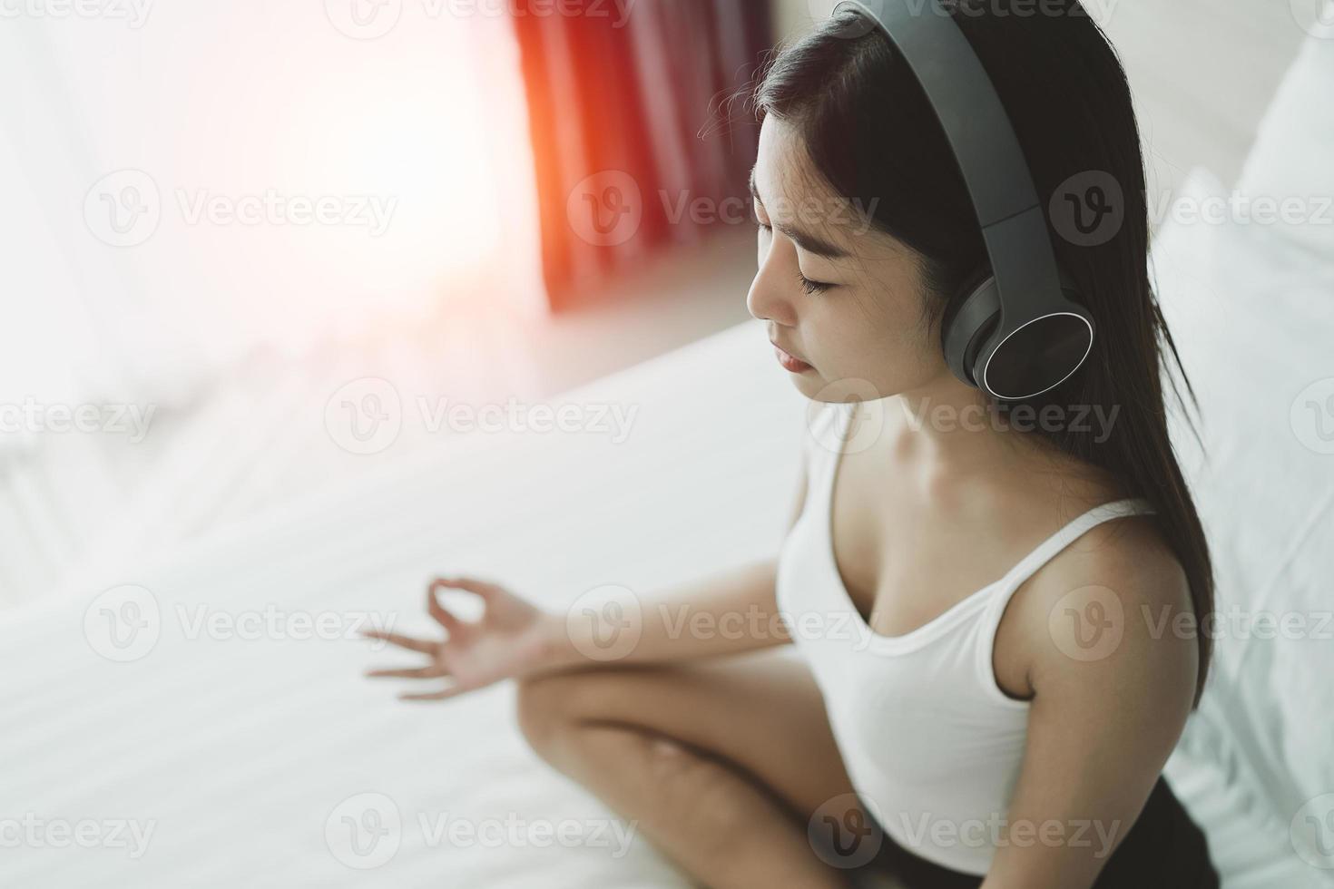 Asian woman wearing headphone practicing yoga lesson, breathing, meditating, doing Ardha Padmasana exercise, Half Lotus pose with mudra gesture, working out. Well being, wellness concept photo