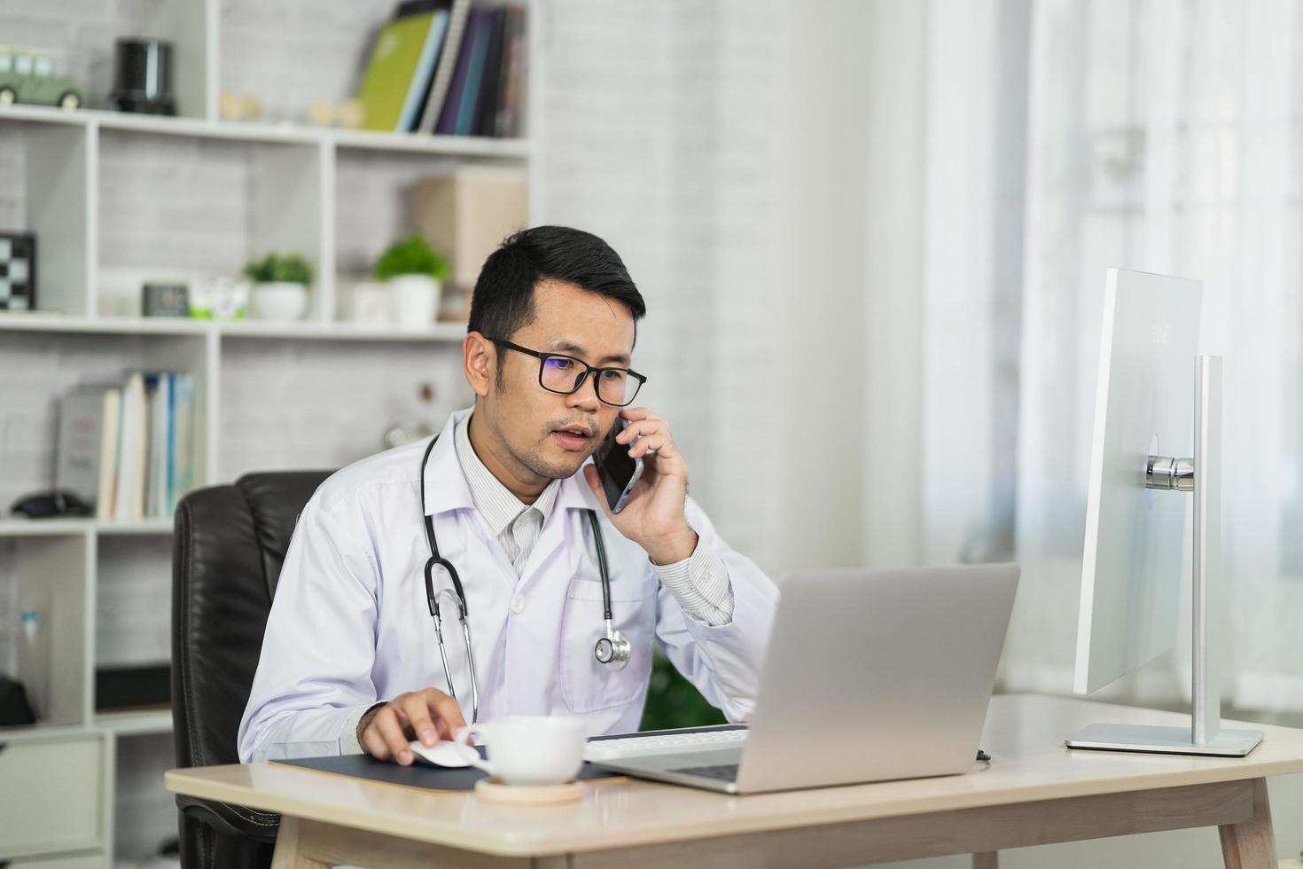 un médico asiático llama a un teléfono mpbile y usa una computadora portátil para consultar al paciente a través de una videollamada en la oficina del hogar. concepto de telemedicina y atención médica foto