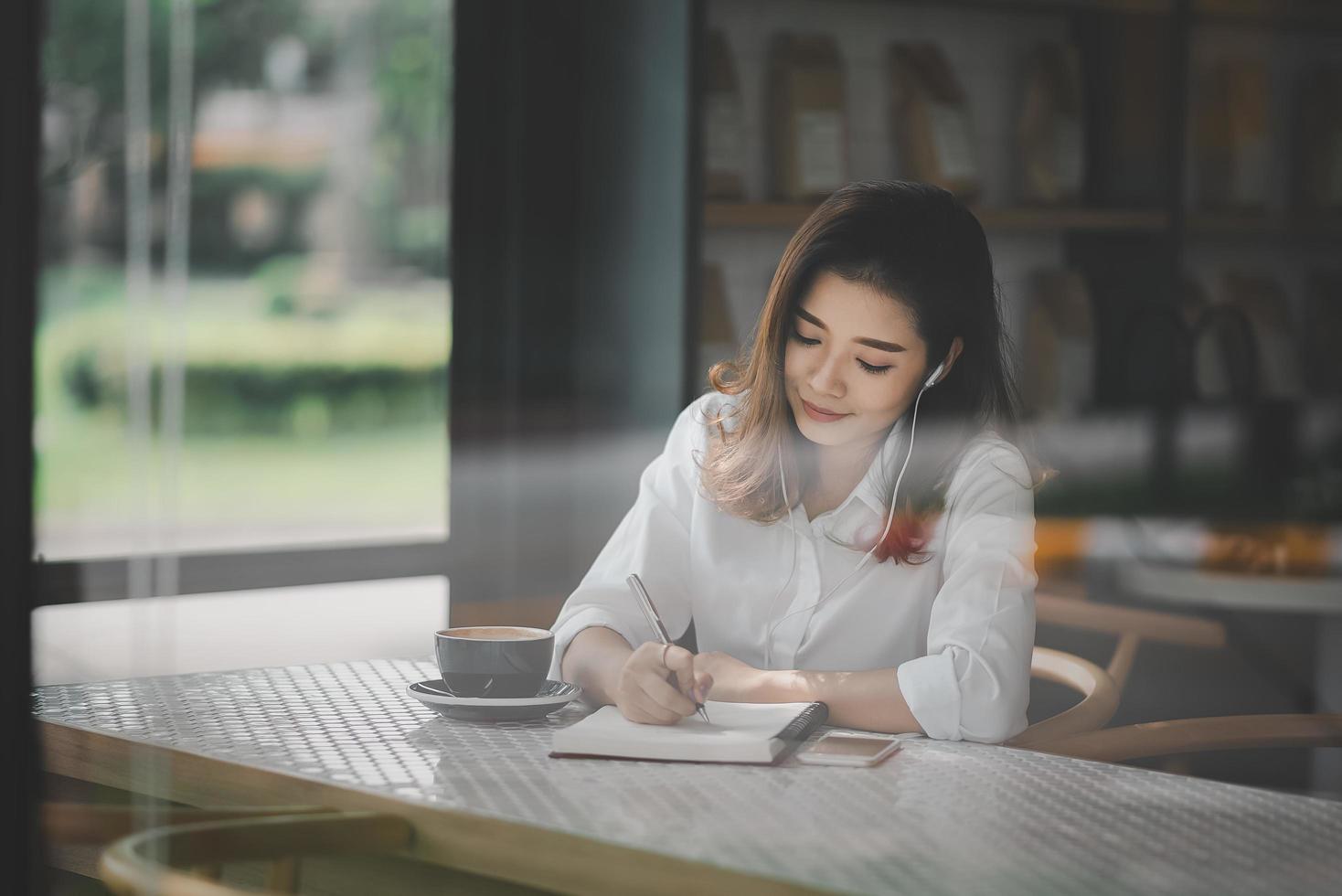 Asian woman sitting and using a pen to write on a paper notebook with a laptop computer and wearing earphones. Work from anywhere concept. Work from home concept. photo