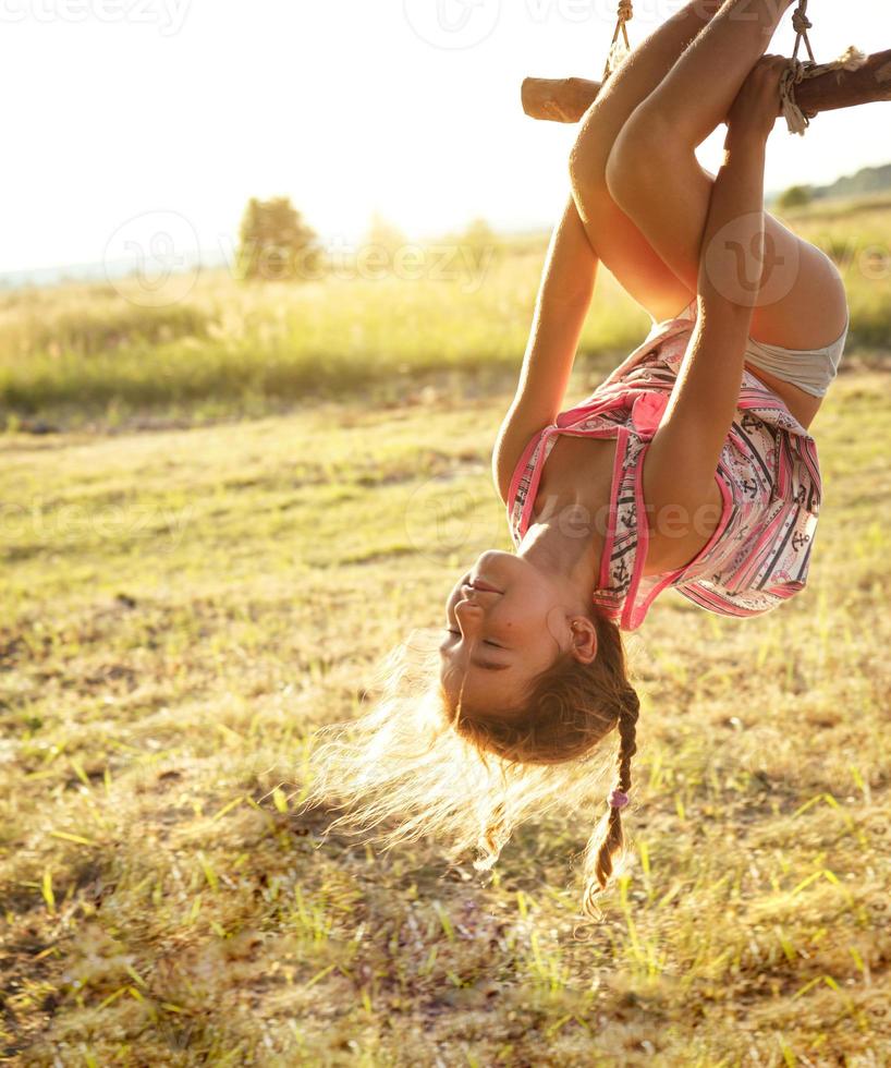 la niña está colgada boca abajo en un árbol en verano a la luz del sol naranja y con un vestido ligero. horario de verano, calor, infancia. retrato divertido con el pelo despeinado foto