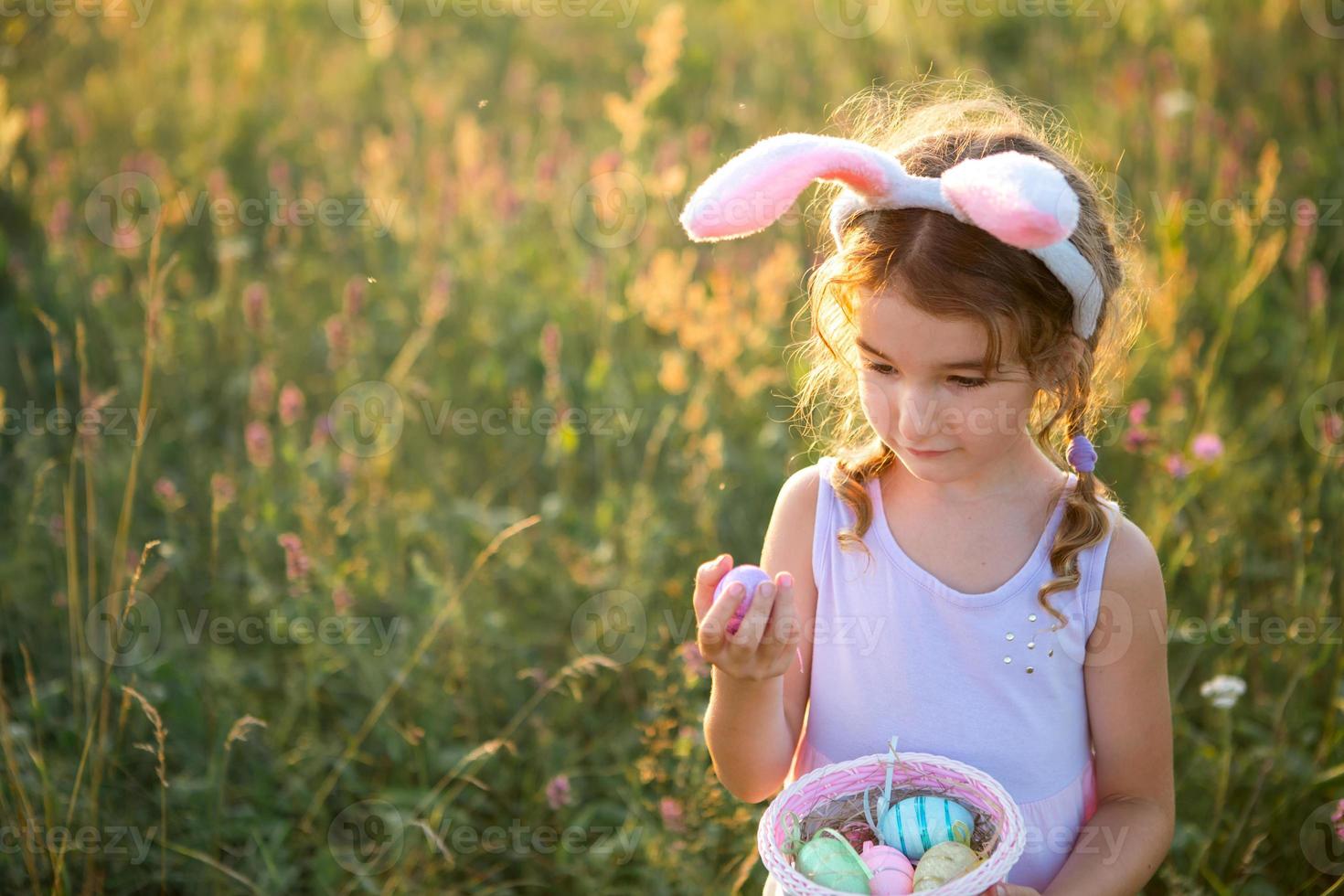 linda chica divertida con huevos de pascua pintados en primavera en la naturaleza en un campo con luz dorada y flores. vacaciones de pascua, conejito de pascua con orejas, huevos coloridos en una canasta. estilo de vida foto