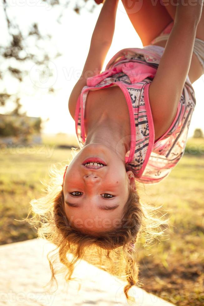 The girl is hanging upside down on a tree in summer in orange sunlight and a light dress. Summer time, heat, childhood. Funny portrait with disheveled hair photo