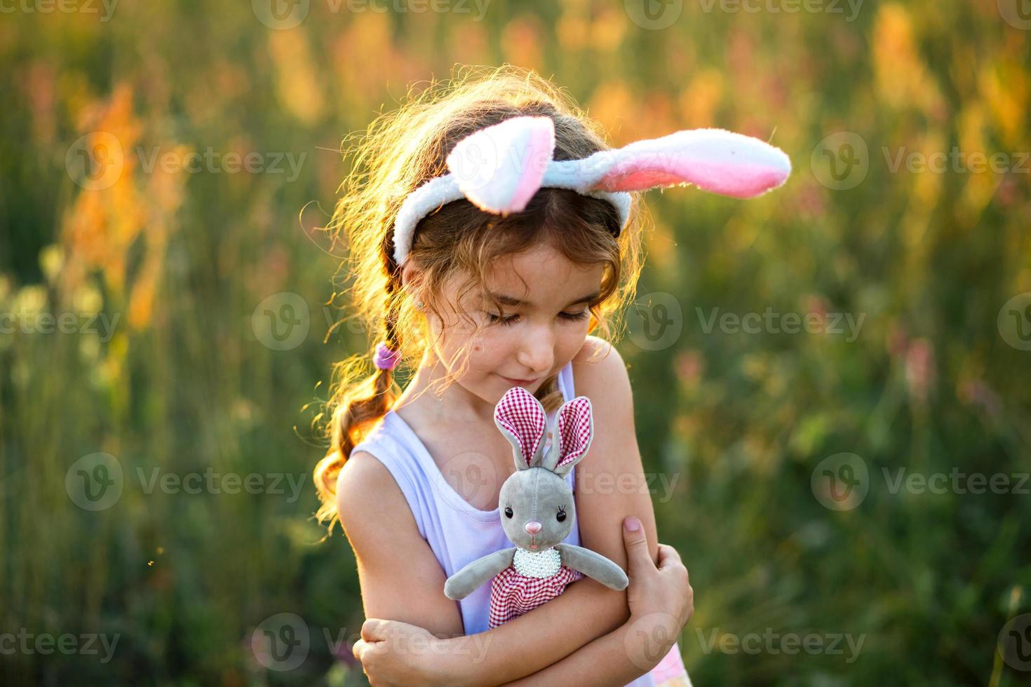 Cute 5-year-old girl with rabbit ears gently hugs a toy rabbit in nature in a blooming field in summer with golden sunlight. Easter, Easter bunny, childhood, happy child, springtime. photo