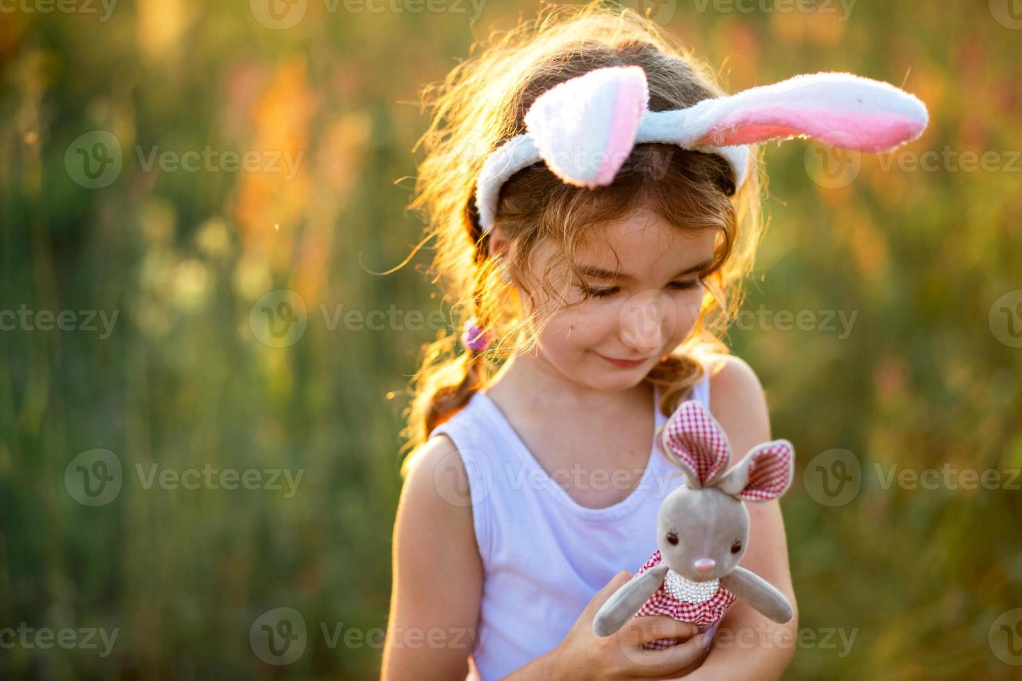 linda niña de 5 años con orejas de conejo abraza suavemente a un conejo de juguete en la naturaleza en un campo floreciente en verano con luz dorada del sol. pascua, conejito de pascua, niñez, niño feliz, primavera. foto