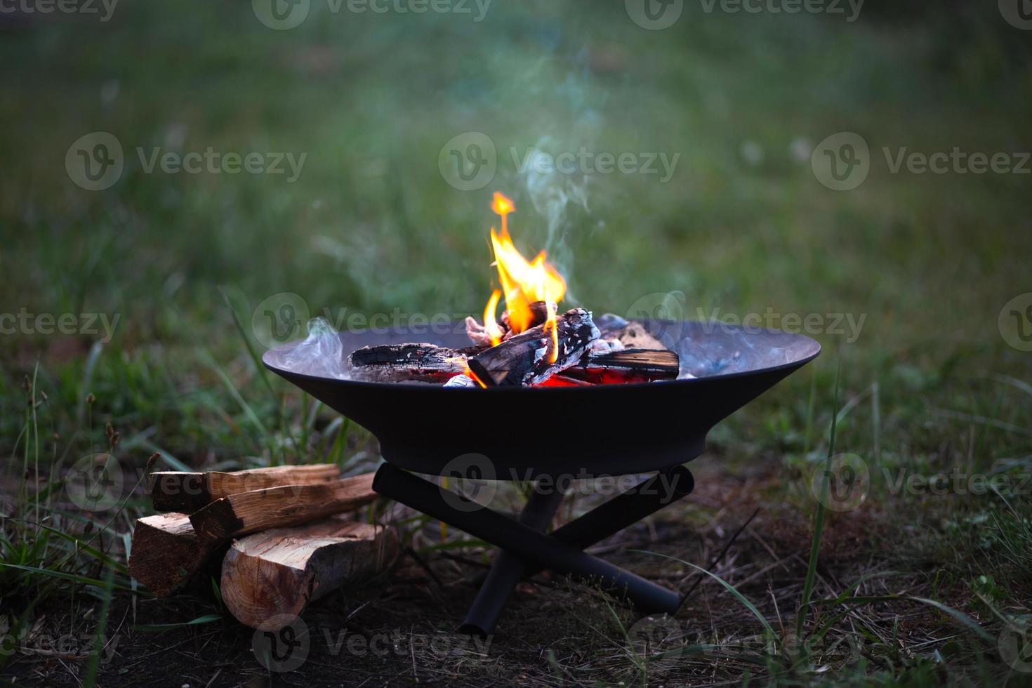 The flame of the fire burns in a metal fire bowl - warm your hands by the fire, stir the firewood with a stick. A hearth with firewood for safe campfire outdoors. Warmth, comfort for gatherings photo