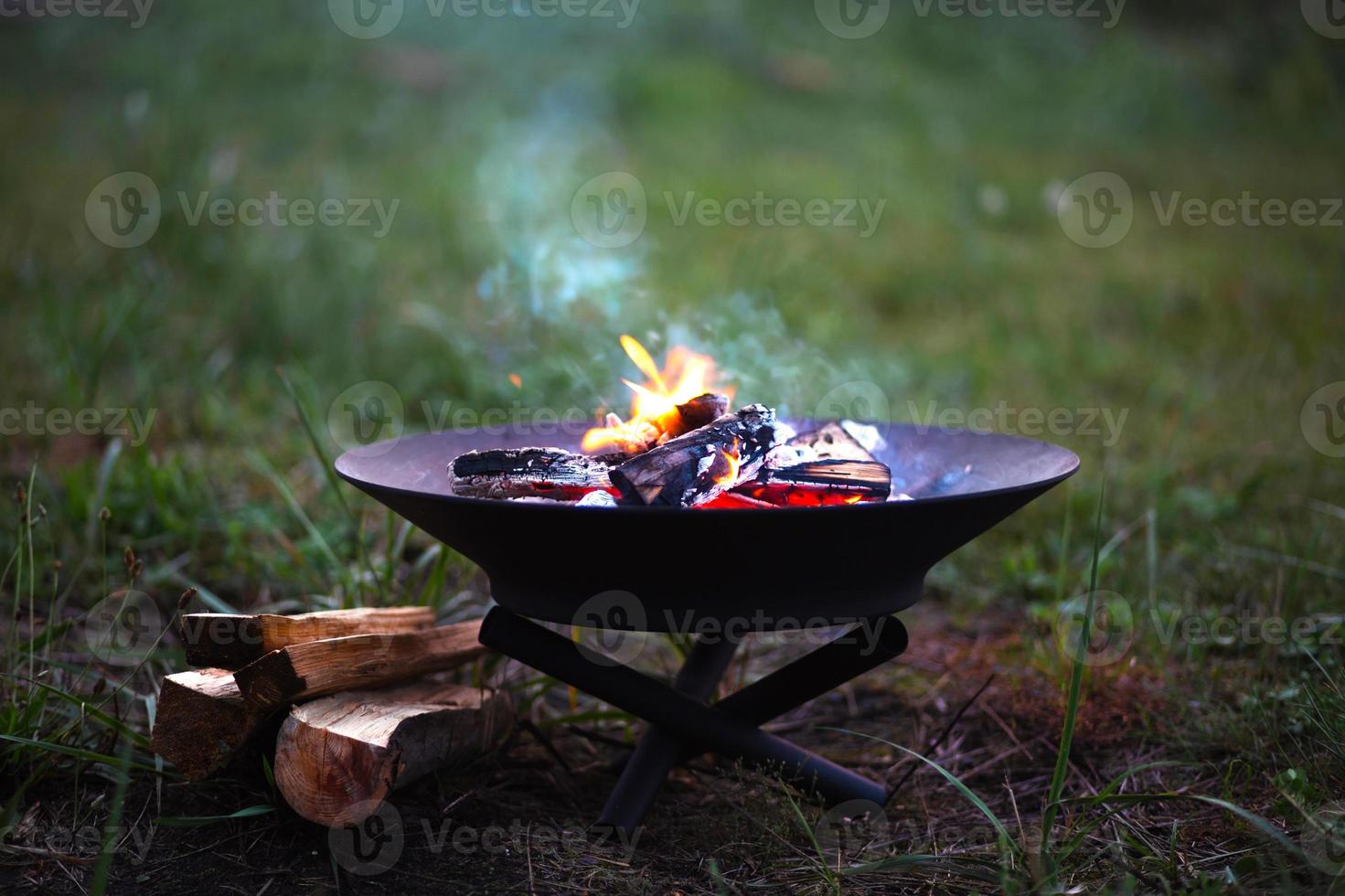 The flame of the fire burns in a metal fire bowl - warm your hands by the fire, stir the firewood with a stick. A hearth with firewood for safe campfire outdoors. Warmth, comfort for gatherings photo