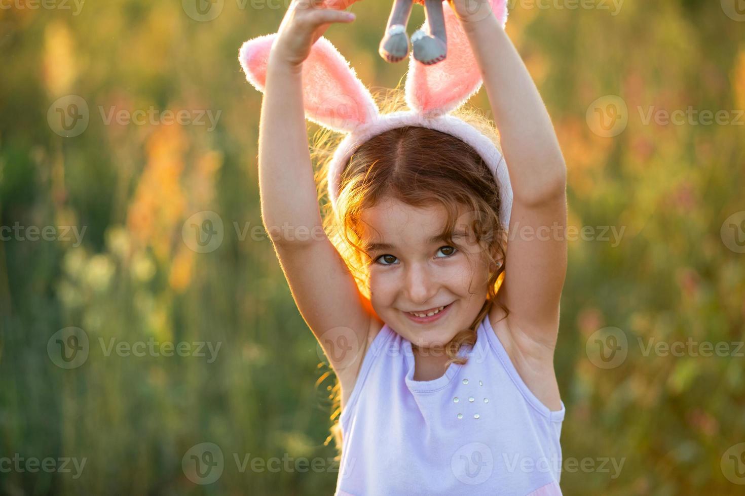 Cute 5-year-old girl with rabbit ears gently hugs a toy rabbit in nature in a blooming field in summer with golden sunlight. Easter, Easter bunny, childhood, happy child, springtime. photo