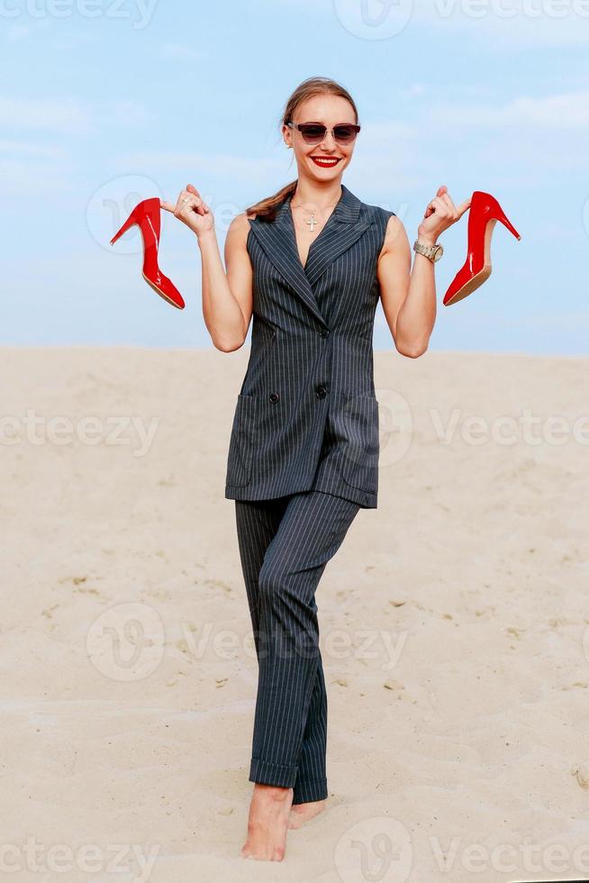 portrait of stylish white young woman in suit, sunglasses, standing with the glossy high heels shoes in her hands outdoor photo