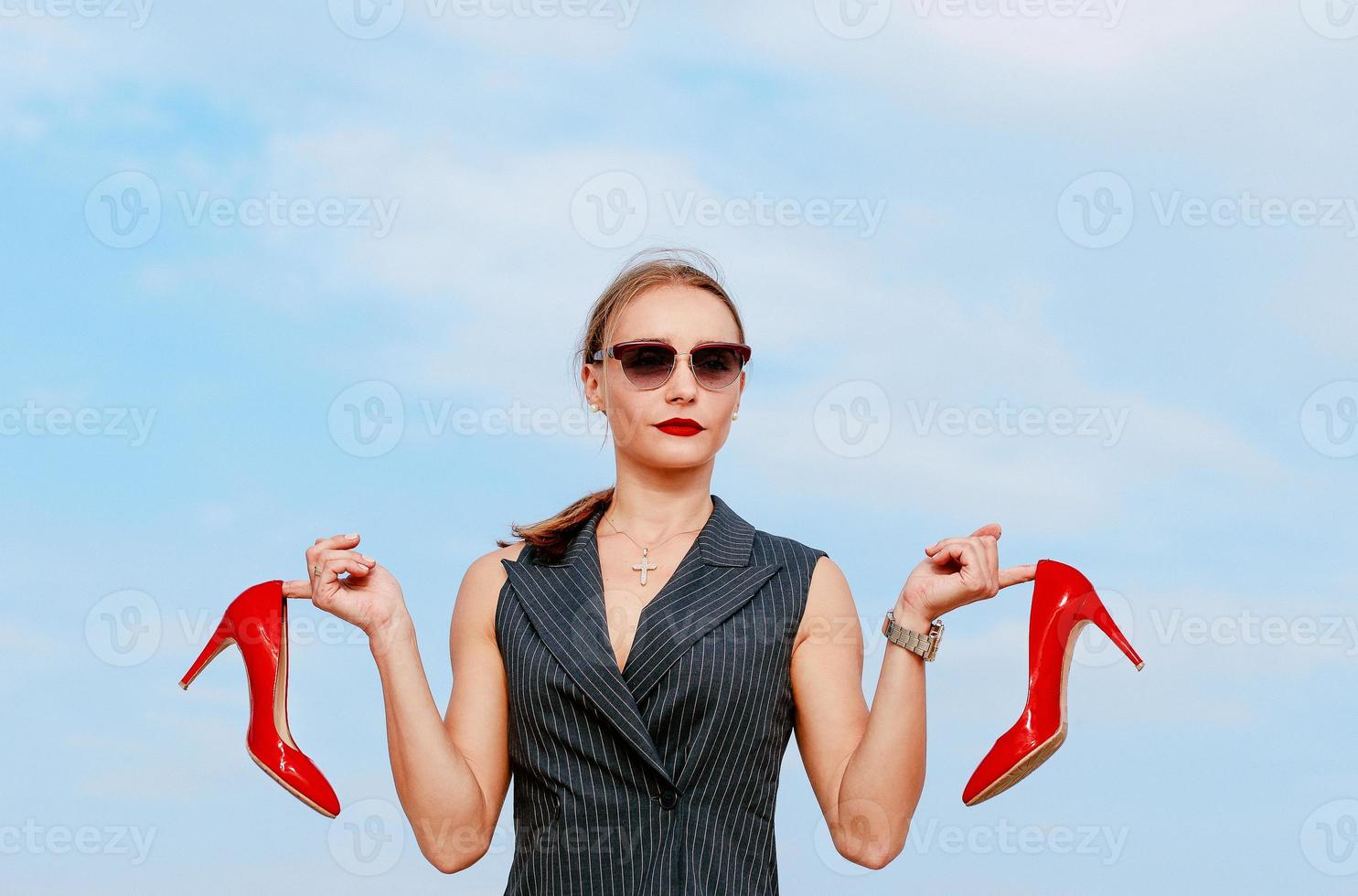 portrait of stylish white young woman in suit, sunglasses, standing with the glossy high heels shoes in her hands outdoor photo