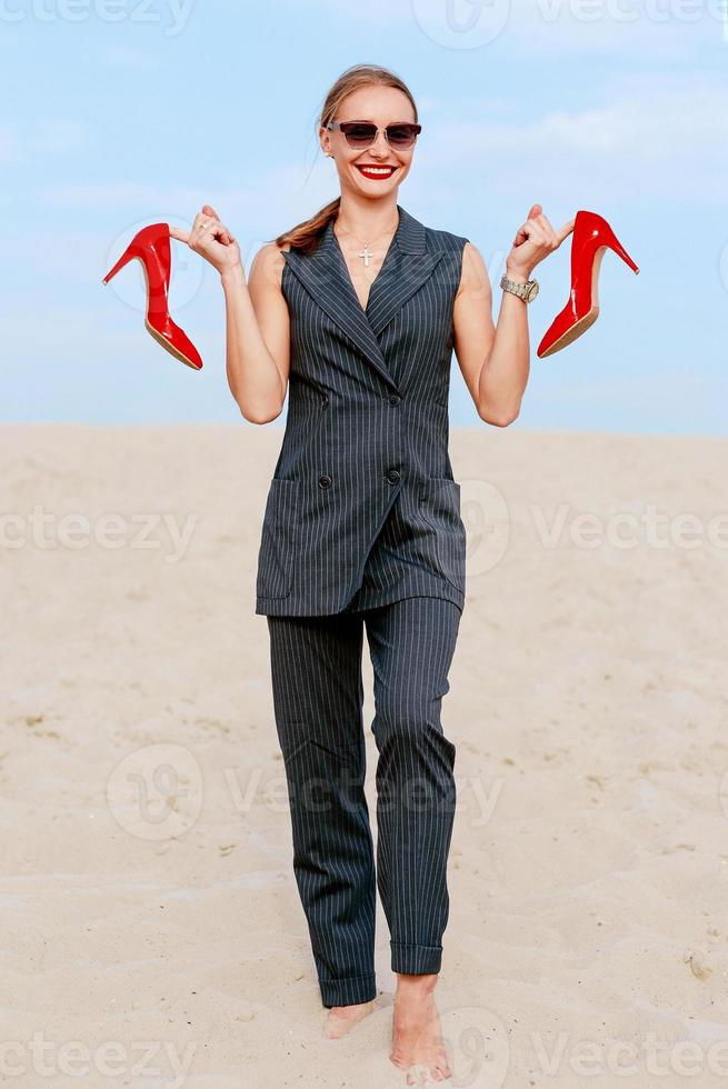 portrait of stylish white young woman in suit, sunglasses, standing with the glossy high heels shoes in her hands outdoor photo