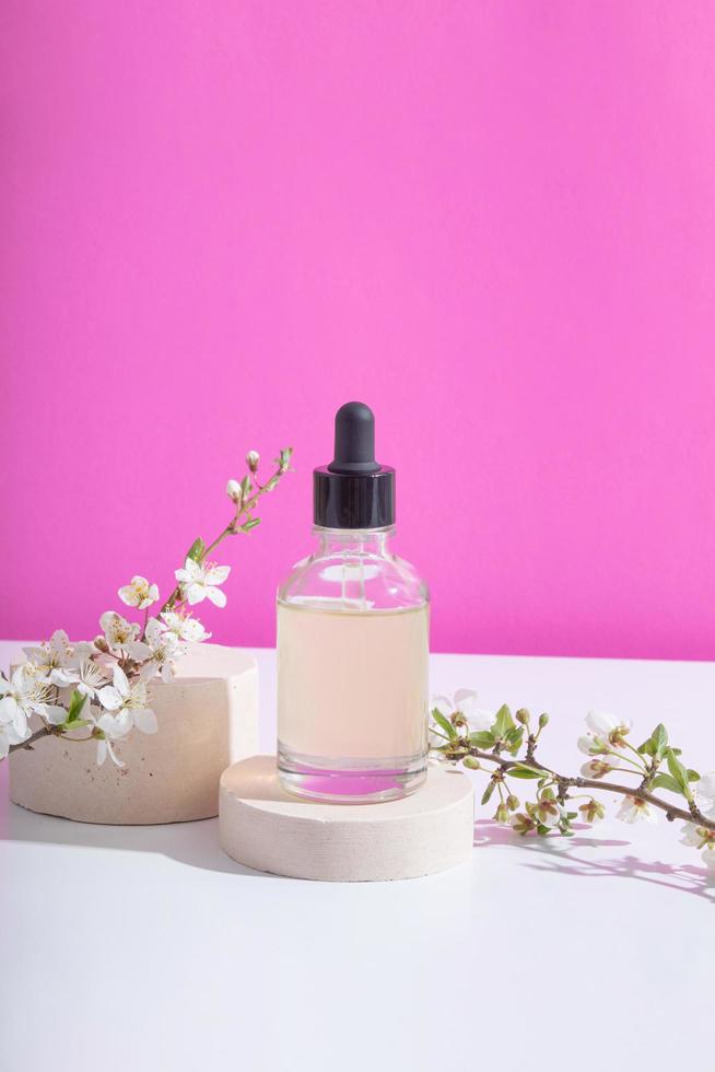 Glass dropper bottle with cosmetic oil on podium with blooming twigs on pink background. Vertical stock photo