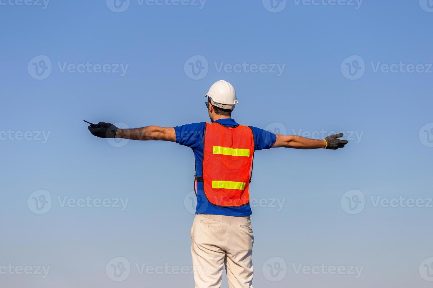 Successful young man with arms wide open, Happy engineer foreman in hardhat and safety vest talks over blue sky background photo