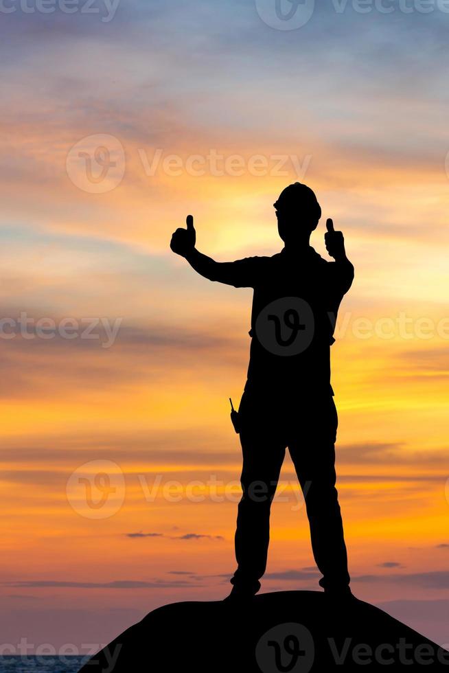 silueta de un trabajador de fábrica con un camino recortado en un sombrero duro con los pulgares hacia arriba, un joven celebra con la mano en el fondo del atardecer foto